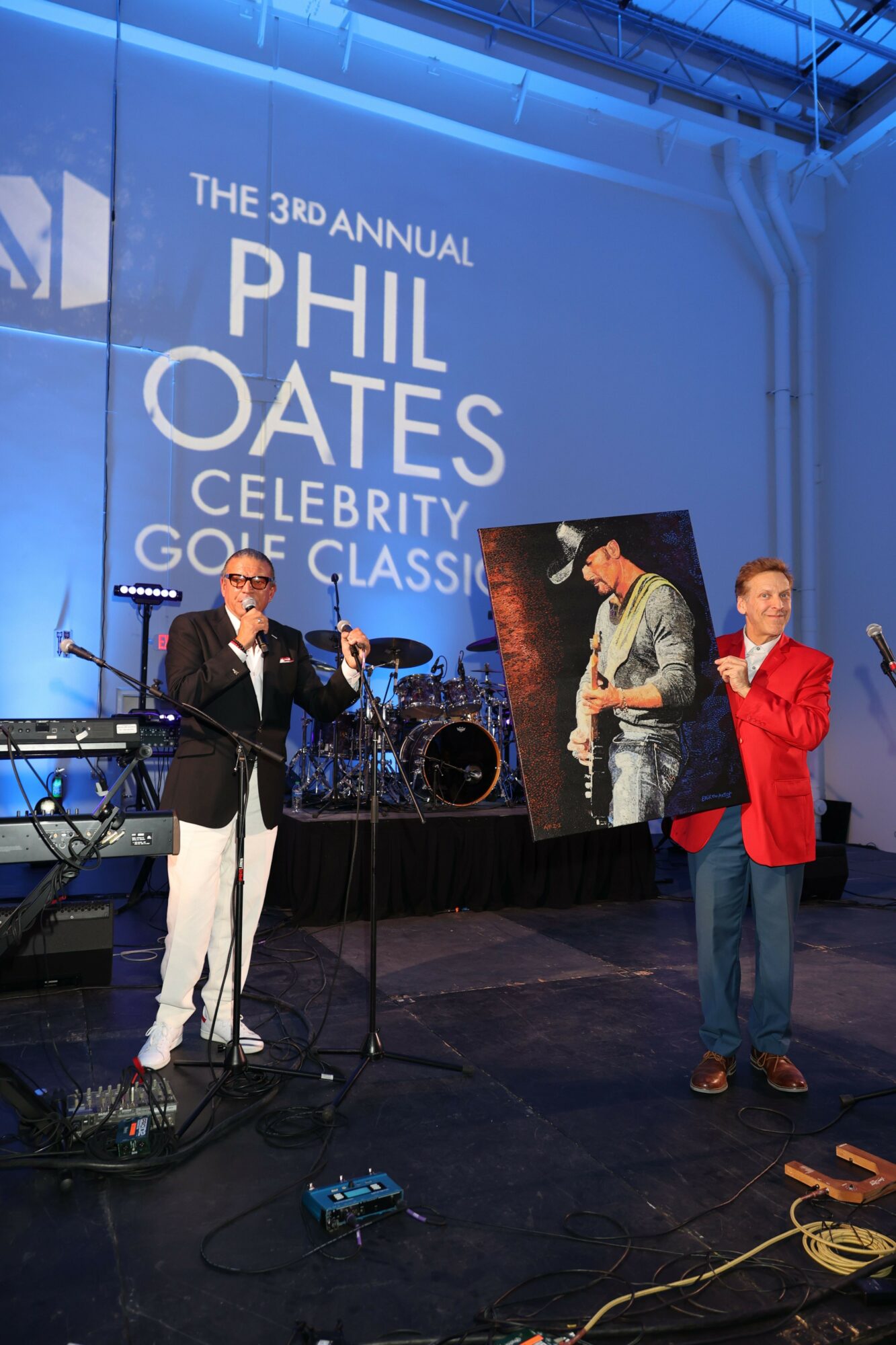 Two men on stage at a podium, one speaking into a microphone, with a large blue backdrop displaying event details.