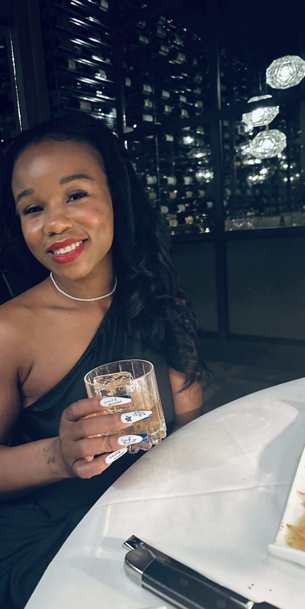 Woman with long dark hair and red lipstick holding a glass of water, sitting at a table with a cityscape background at night.