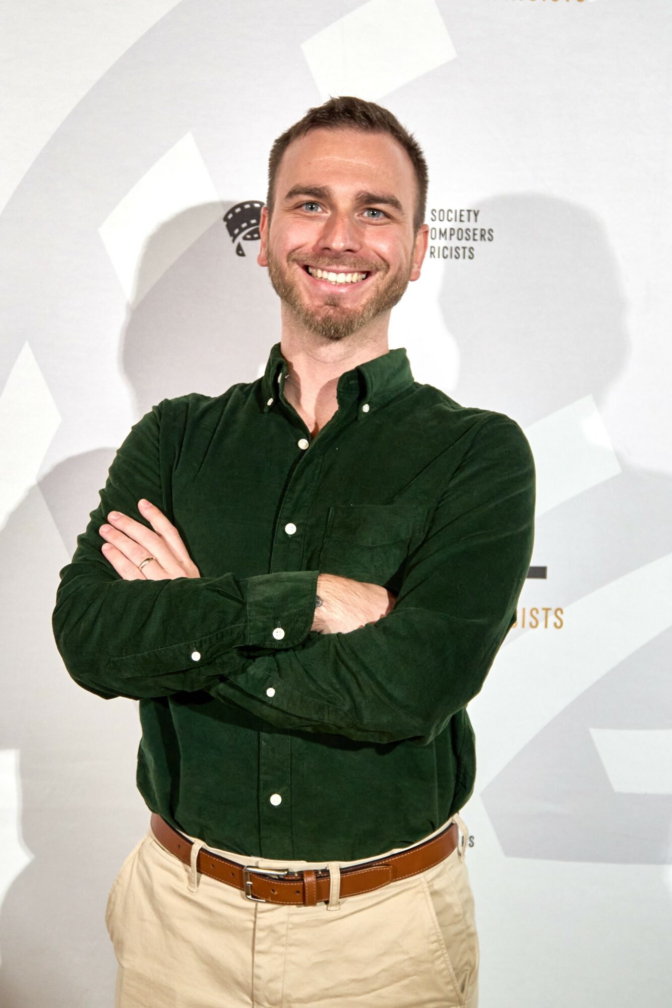 Smiling man with crossed arms wearing a dark green shirt and beige pants, standing in front of a light background.