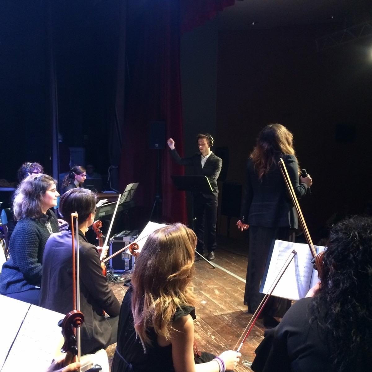 Orchestra conductor leading musicians on stage during a performance, with a woman holding a violin and music stands visible.