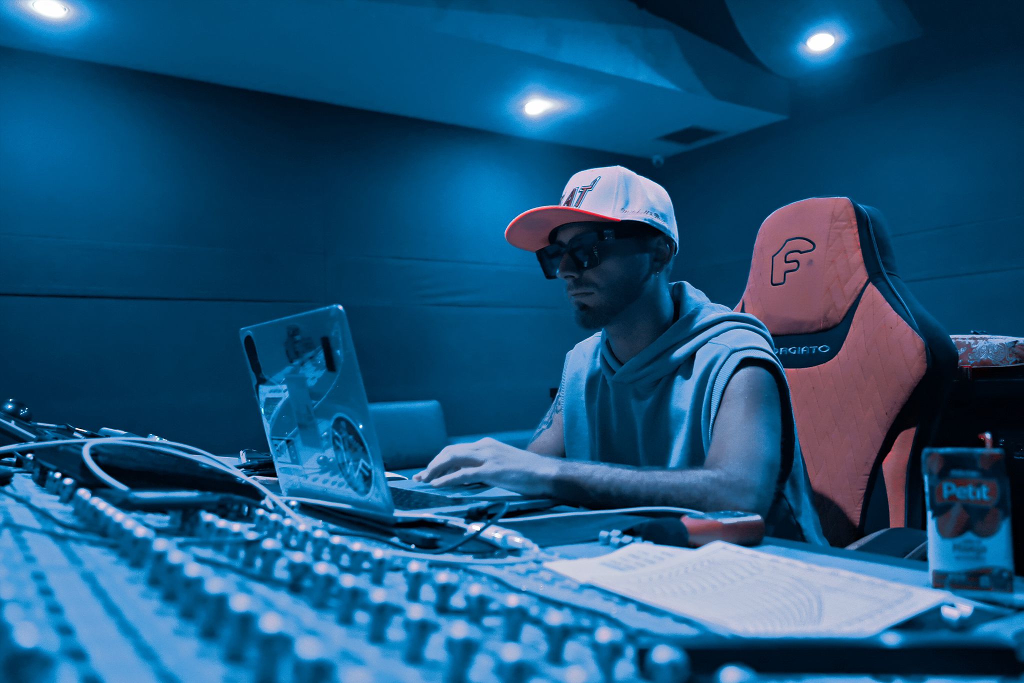 Person wearing a cap and sunglasses working on a laptop in a dimly lit room with audio equipment.