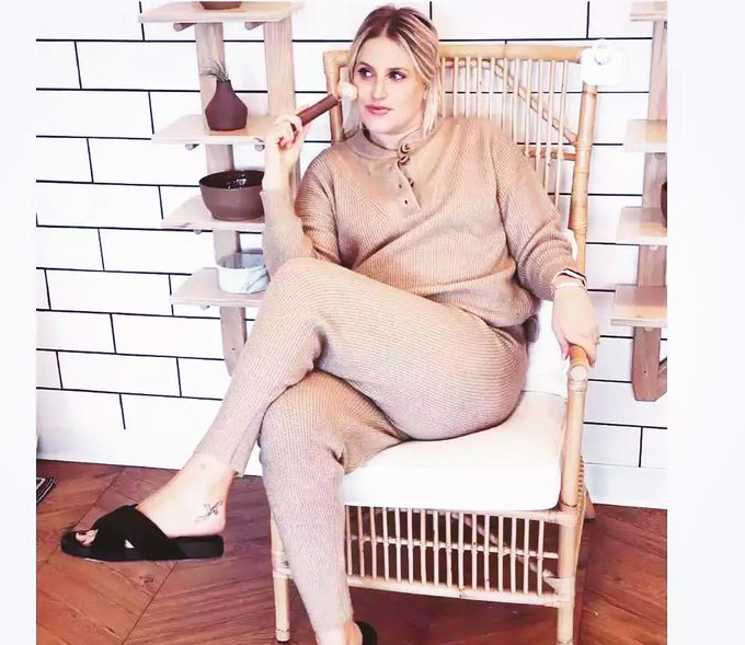 Woman sitting on a chair with a white cushion, in a room with white tiled wall, wearing beige outfit, holding a phone.