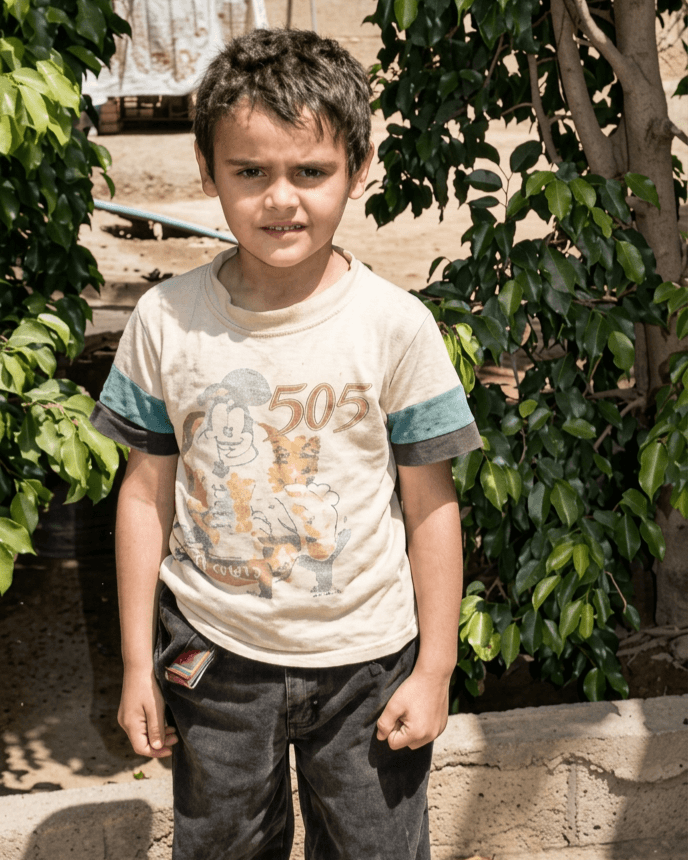 Young boy with short dark hair standing outdoors among green bushes, wearing a light-colored T-shirt and dark pants.