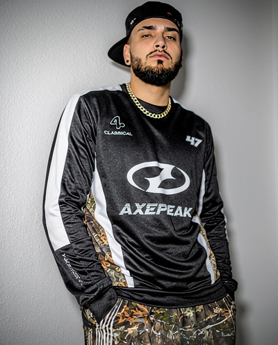 Young man with a beard and cap, wearing a black sports jersey and camouflage pants, standing against a gray wall.