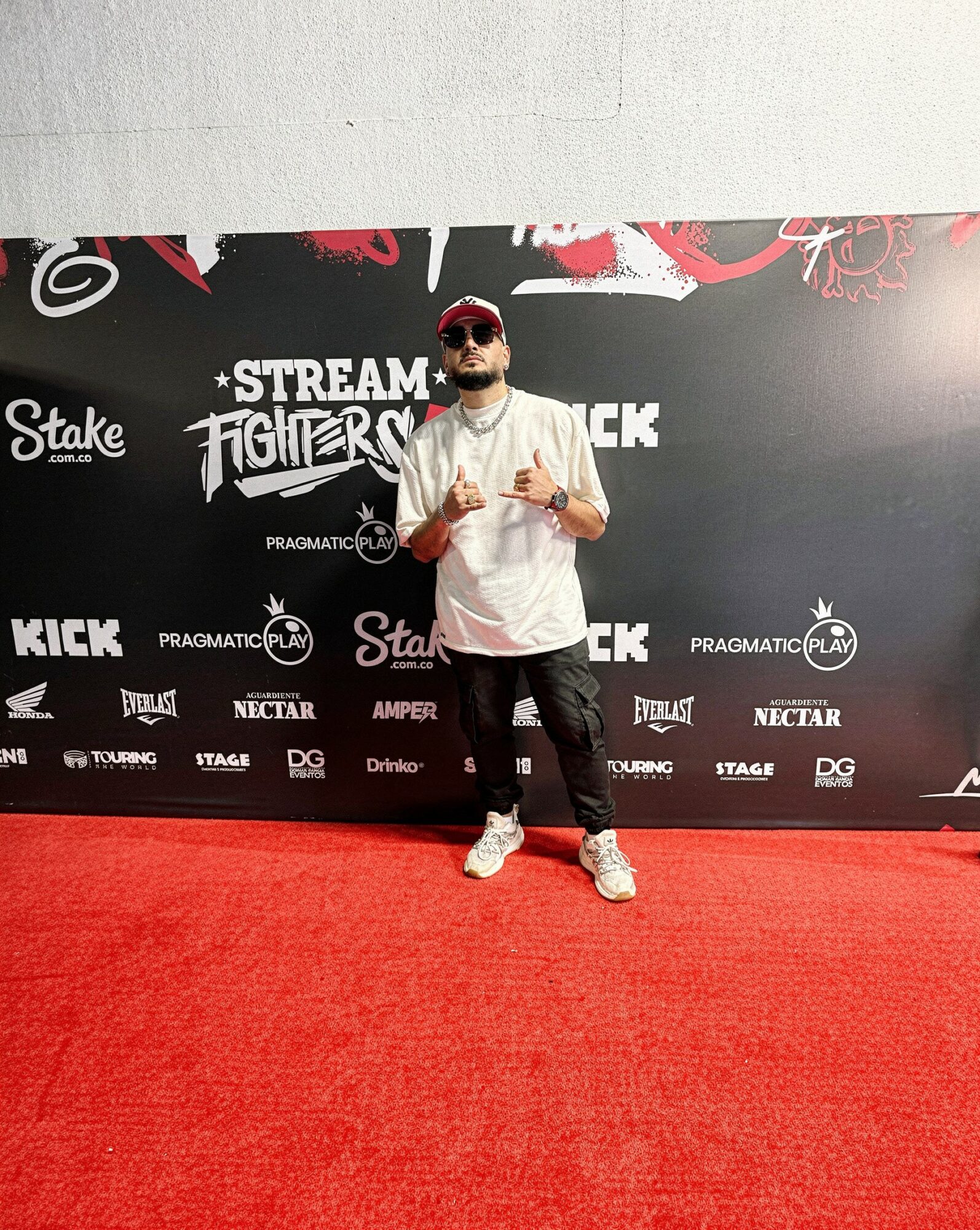 Man with sunglasses and a beard standing on red carpet in front of a black backdrop with logos and text.