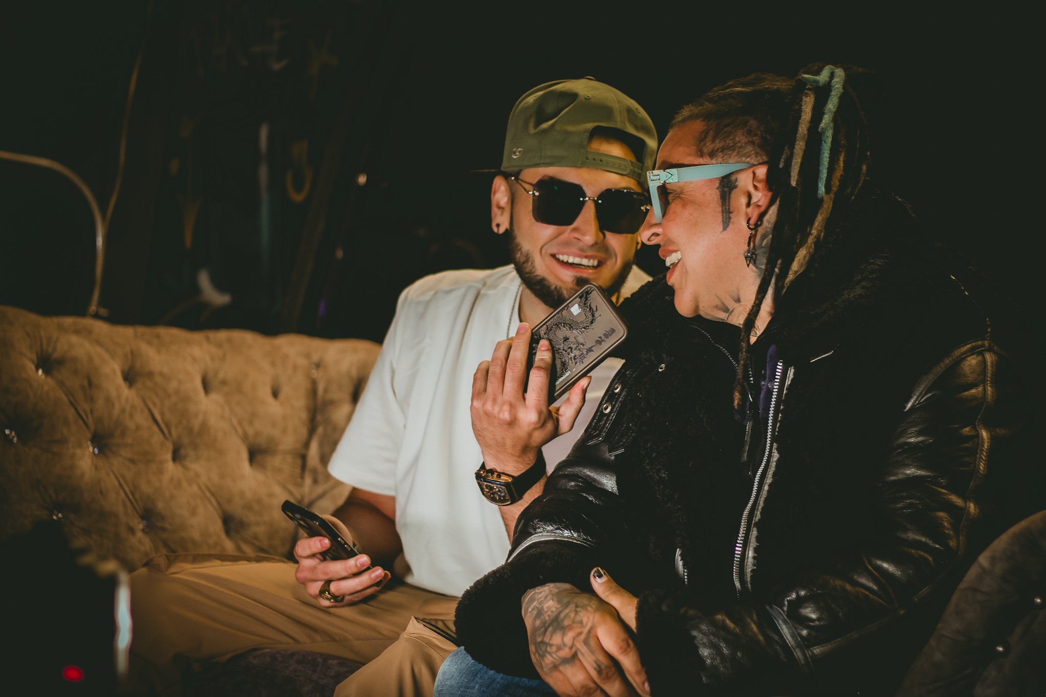 Two men sitting on a couch, smiling and looking at a phone together, in a dark setting.