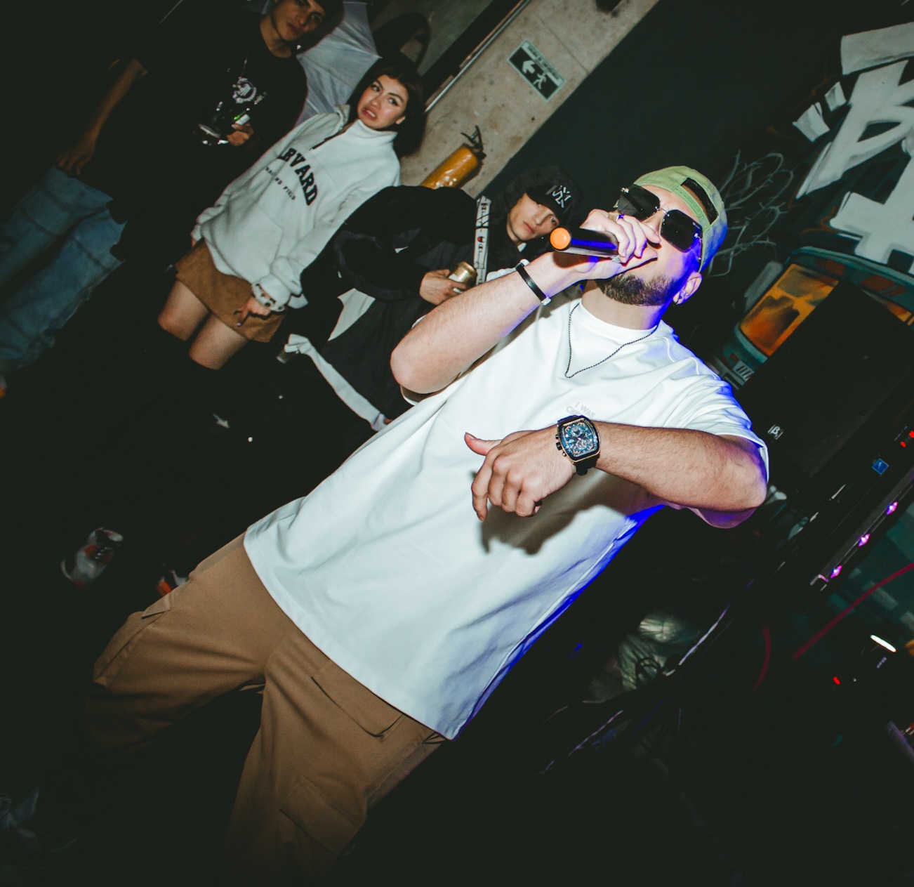 Man wearing sunglasses and a cap, holding a microphone, performing on stage with a group of people in the background.