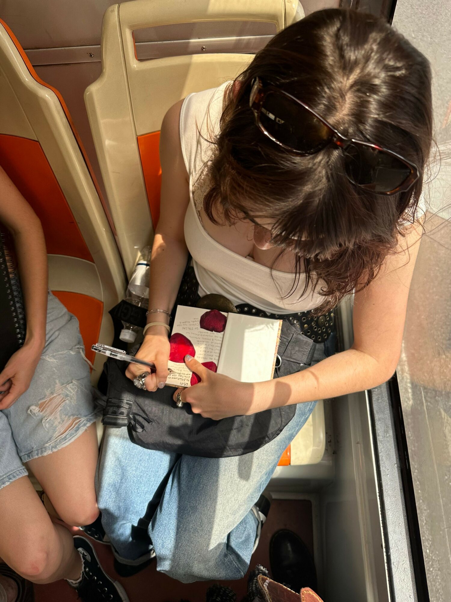 Person with dark hair, wearing sunglasses, white shirt, and ripped jeans, sitting on a bus seat, holding a small object.
