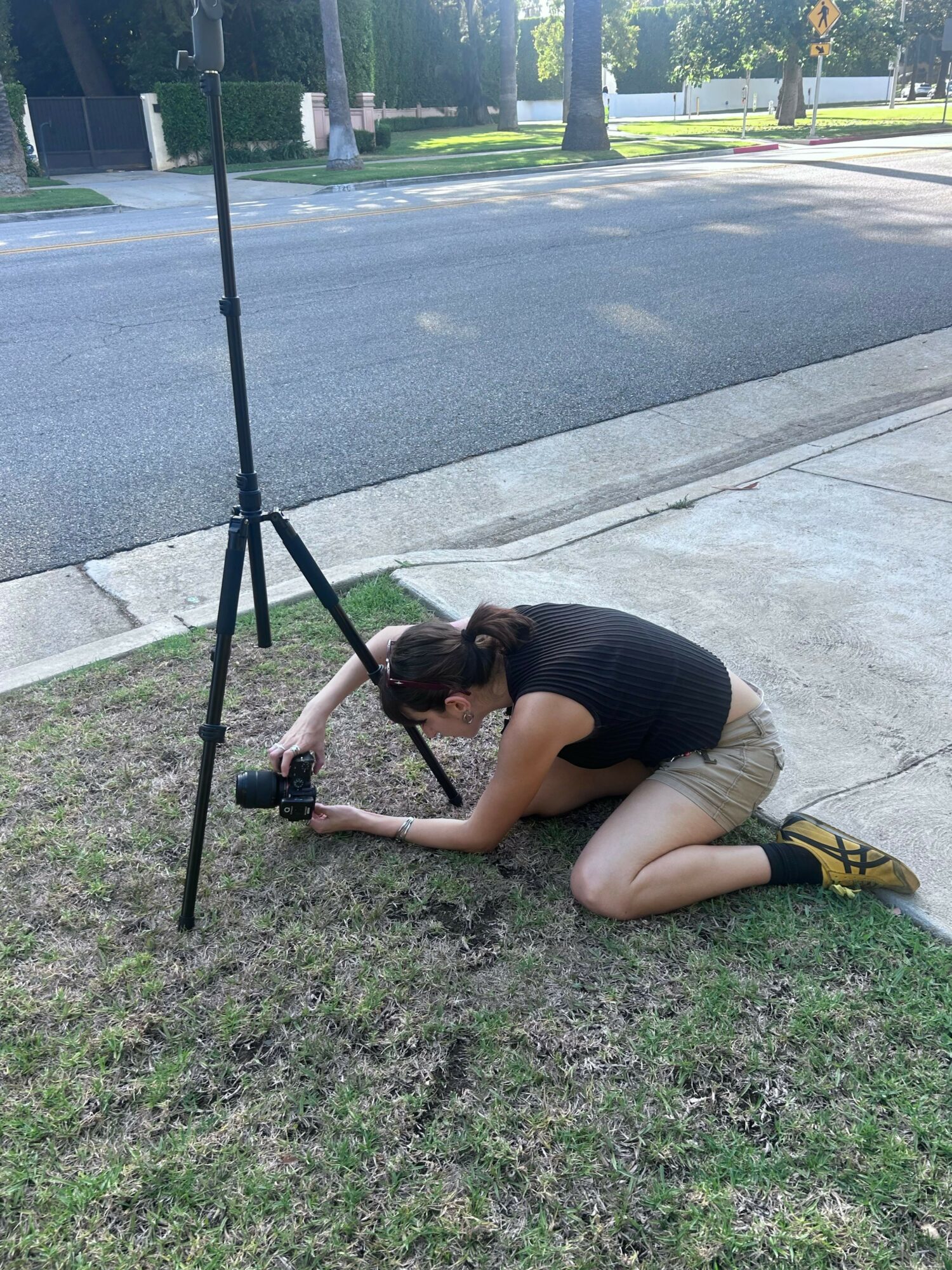 Person lying on grass next to sidewalk, holding a camera, with a pole nearby, in a park-like setting.