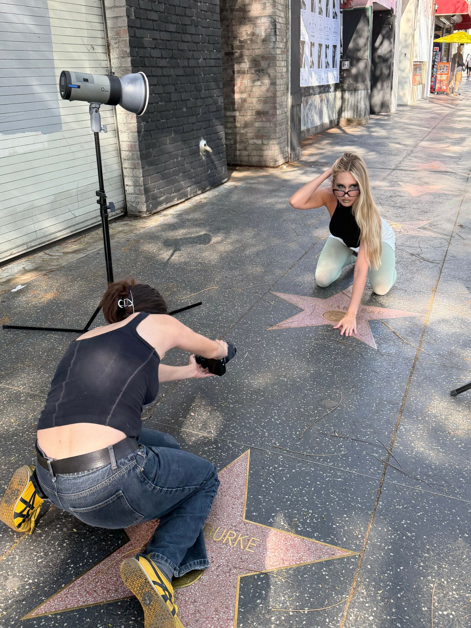 Person crouching on Hollywood Walk of Fame star, taking a photo of woman posing nearby, sidewalk scene, sunny day.
