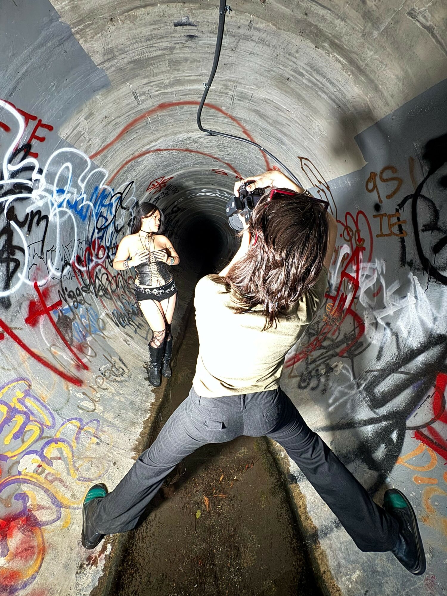 Person with long hair and headphones leaning inside a graffiti-covered tunnel, holding a spray paint can, with graffiti on the walls.