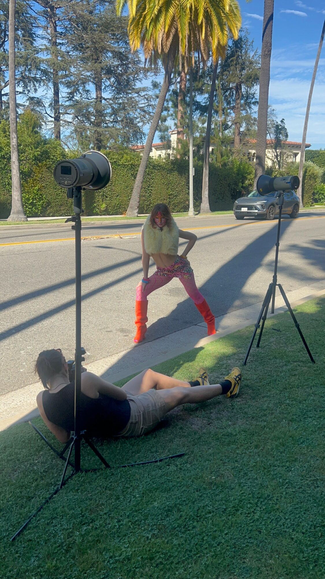 Person lying on grass with photography equipment, woman standing on sidewalk, trees and parked cars in background.