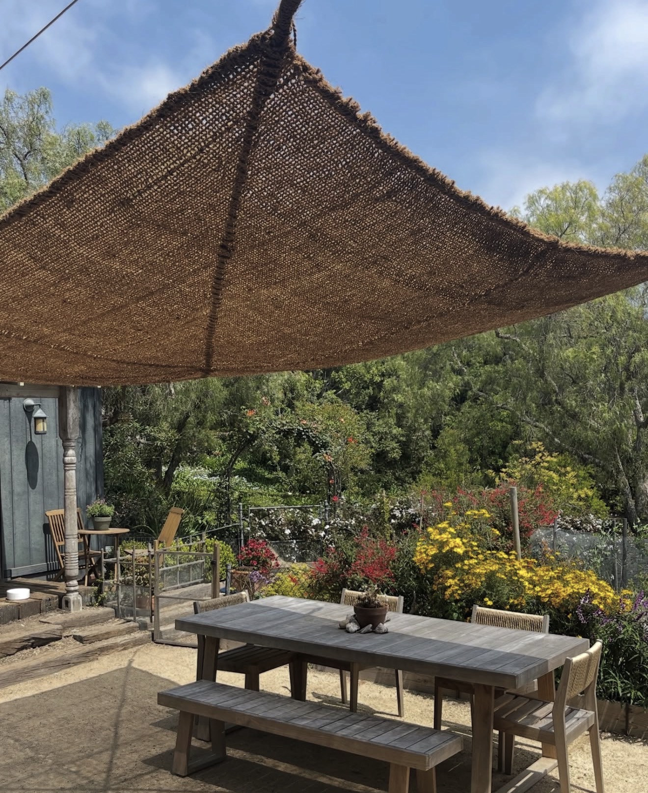 Outdoor patio with wooden table and benches, surrounded by colorful flowers and trees, under a large shade sail.