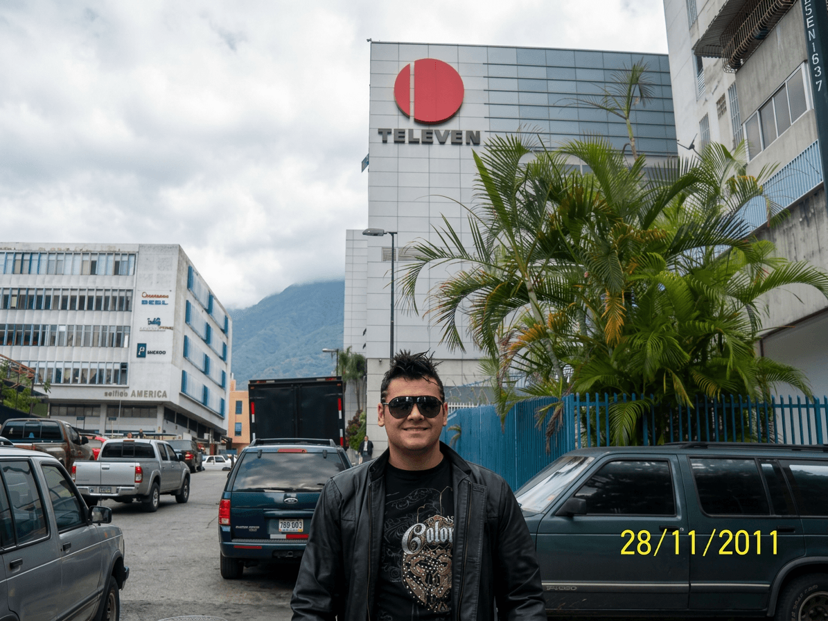 Young man wearing sunglasses and a black jacket standing in a parking lot with buildings and a palm tree behind him.