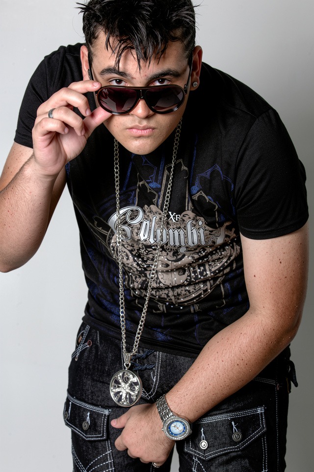 Young man with dark hair wearing sunglasses, black t-shirt, and jewelry, looking at camera, against plain background.
