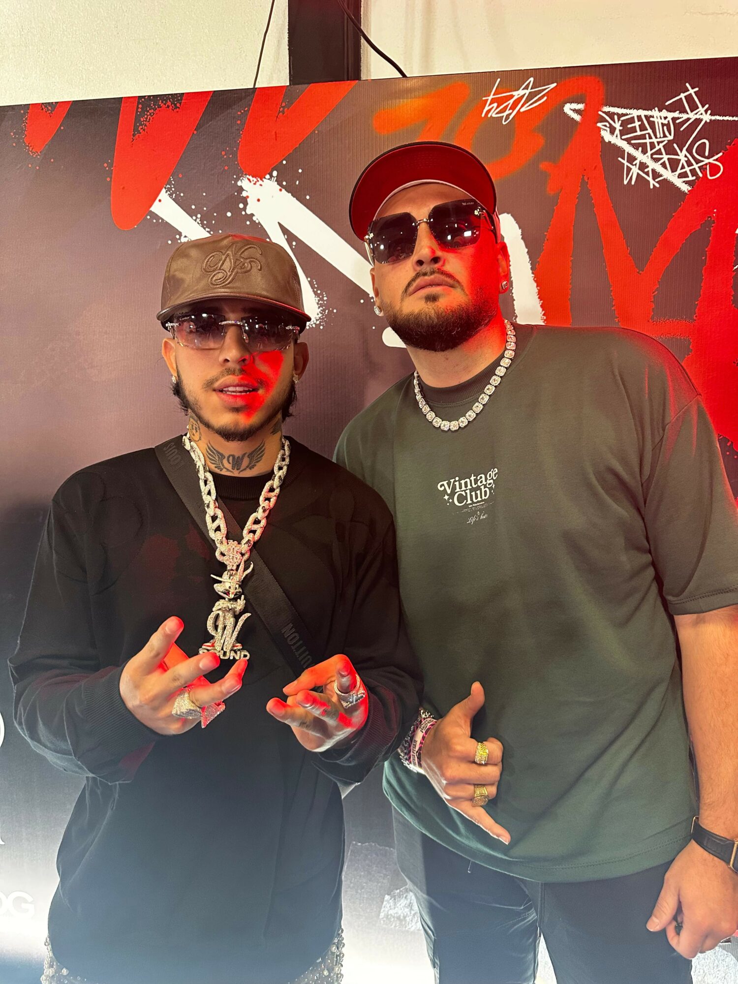 Two men wearing sunglasses and casual clothing pose in front of a red and black background with graffiti-style text.