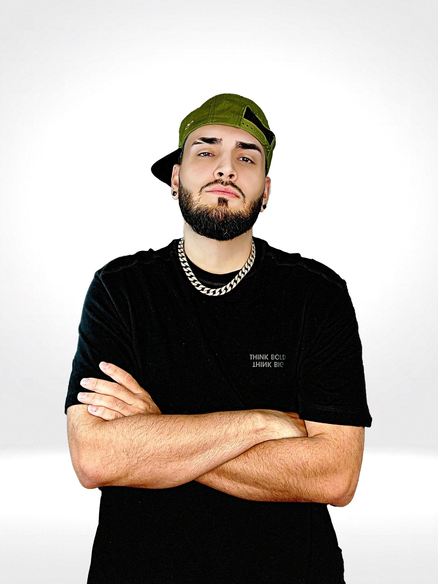 Young man with a beard, wearing a black shirt, green cap backward, and a chain necklace, crossing arms, against a white background.