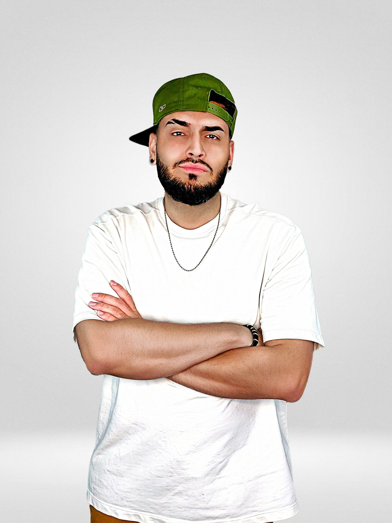 Young man with crossed arms, wearing a green cap backward and a white t-shirt, standing against a plain background.