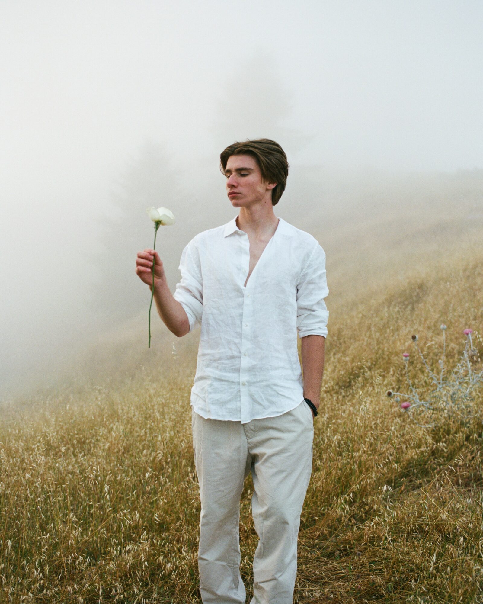 Young man standing in a foggy field holding a white flower, wearing a white shirt and light-colored pants.
