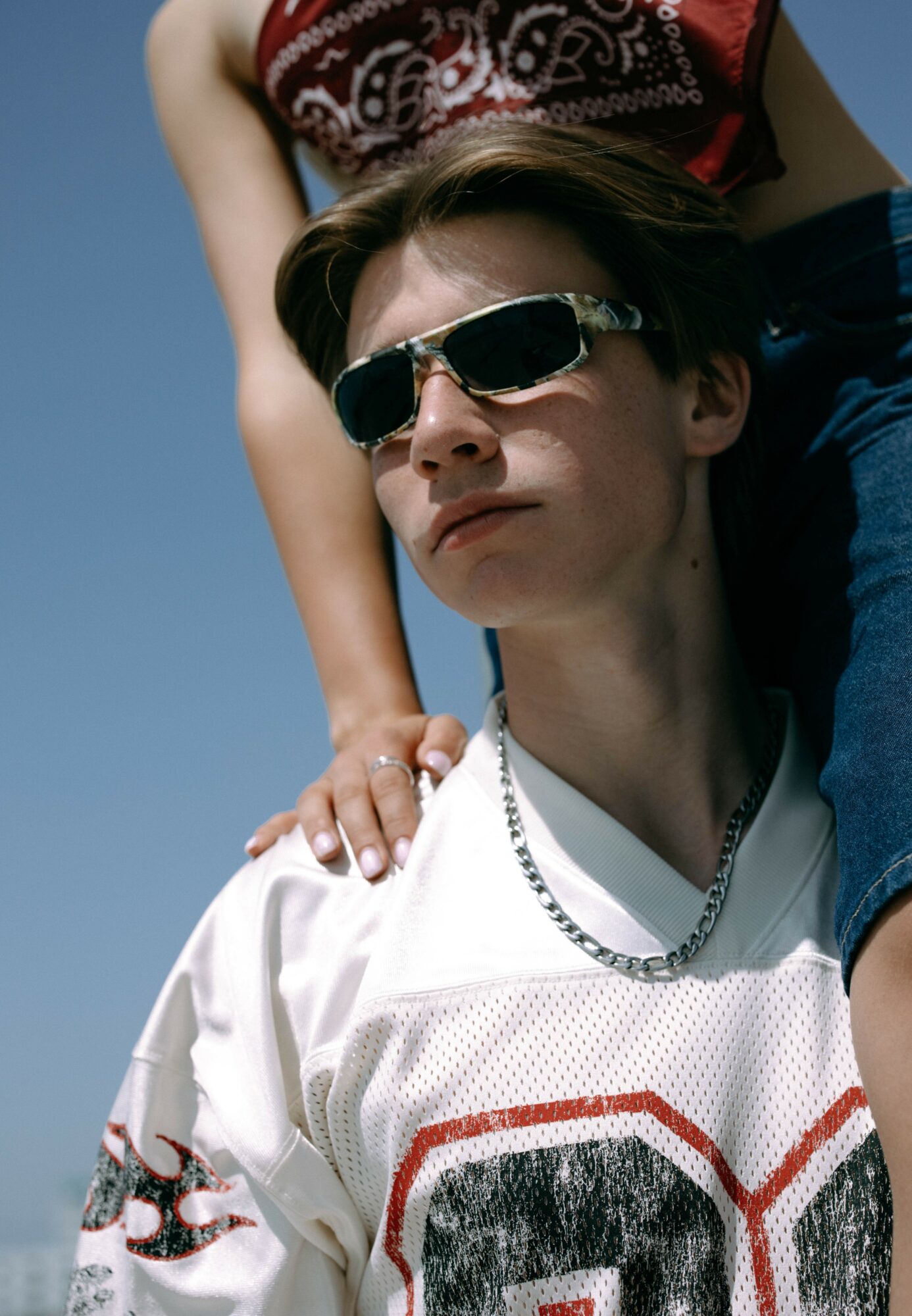 Young person wearing sunglasses and a white sports jersey, with another person resting their hand on their shoulder, against a clear blue sky.