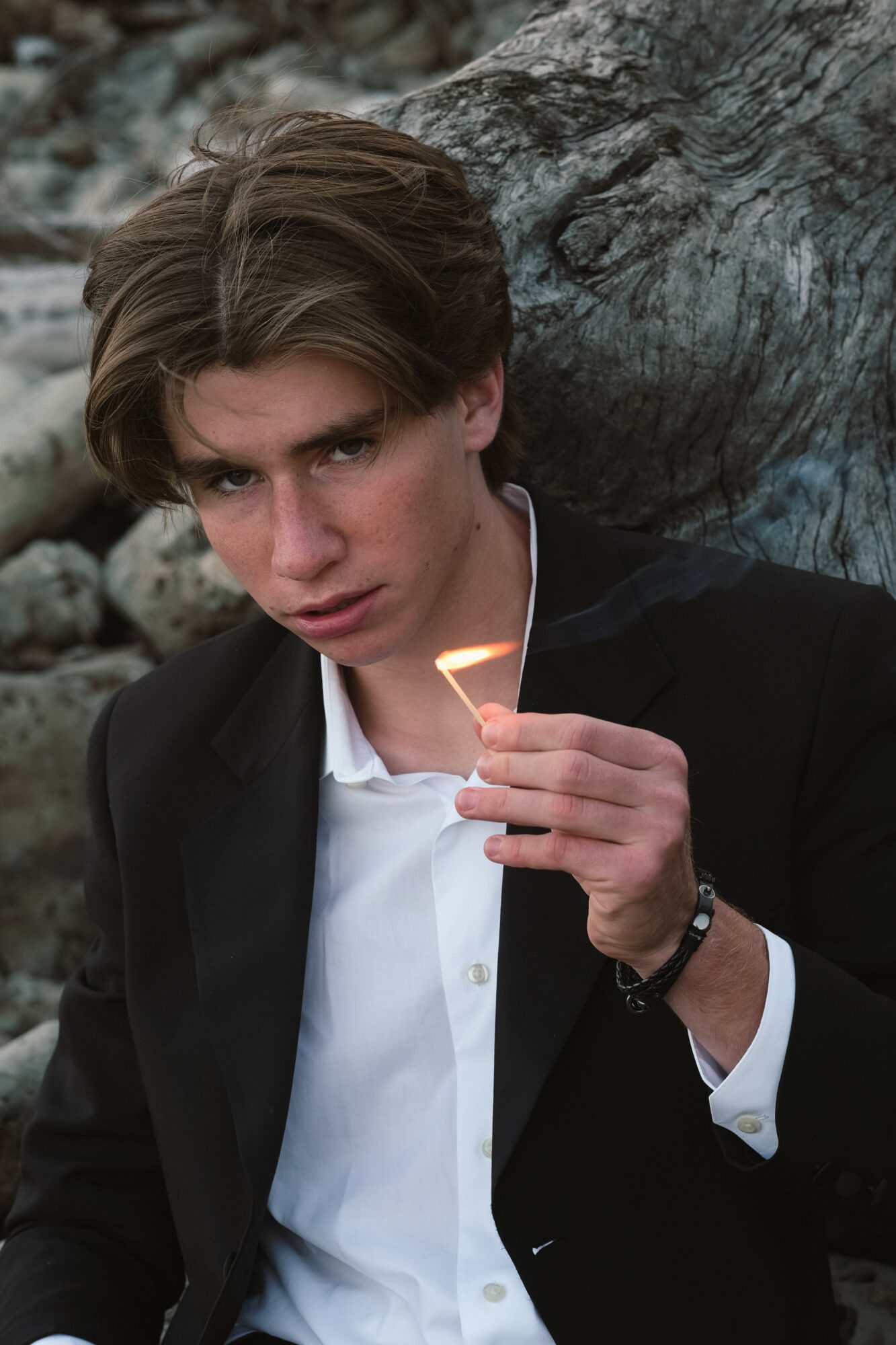 Young man with brown hair in a black suit and white shirt holding a lit match outdoors near rocks.