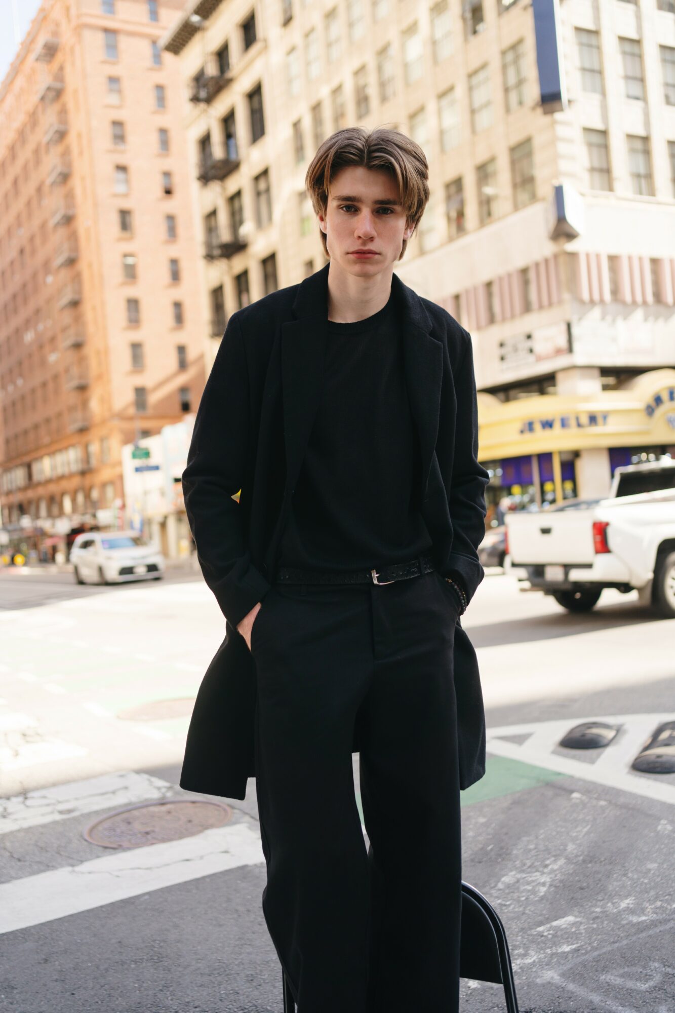 Young man with dark hair and black coat standing on city street with tall buildings and cars in background.