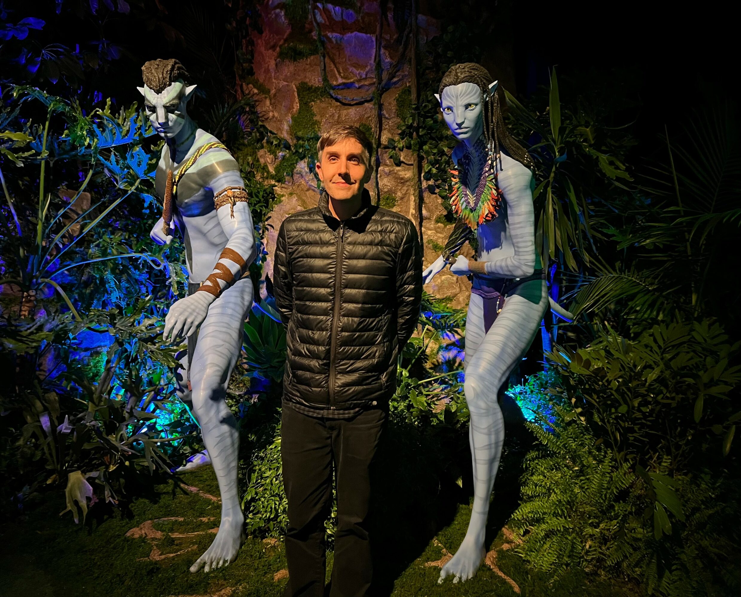 Person standing between two tall, blue-skinned, alien-like figures in a lush, colorful jungle setting at night.
