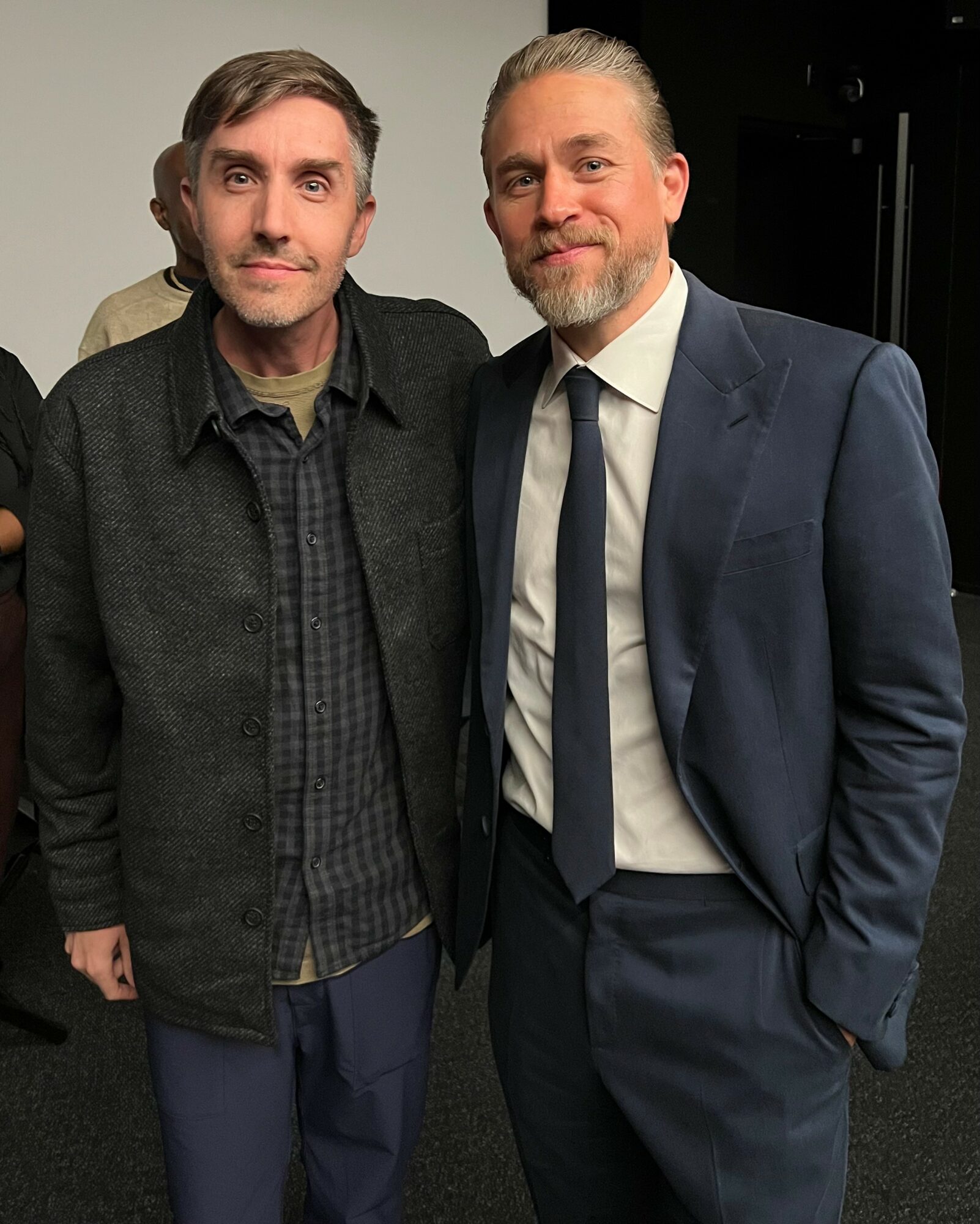 Two men stand side by side, one in casual clothing and the other in a suit, smiling at the camera.