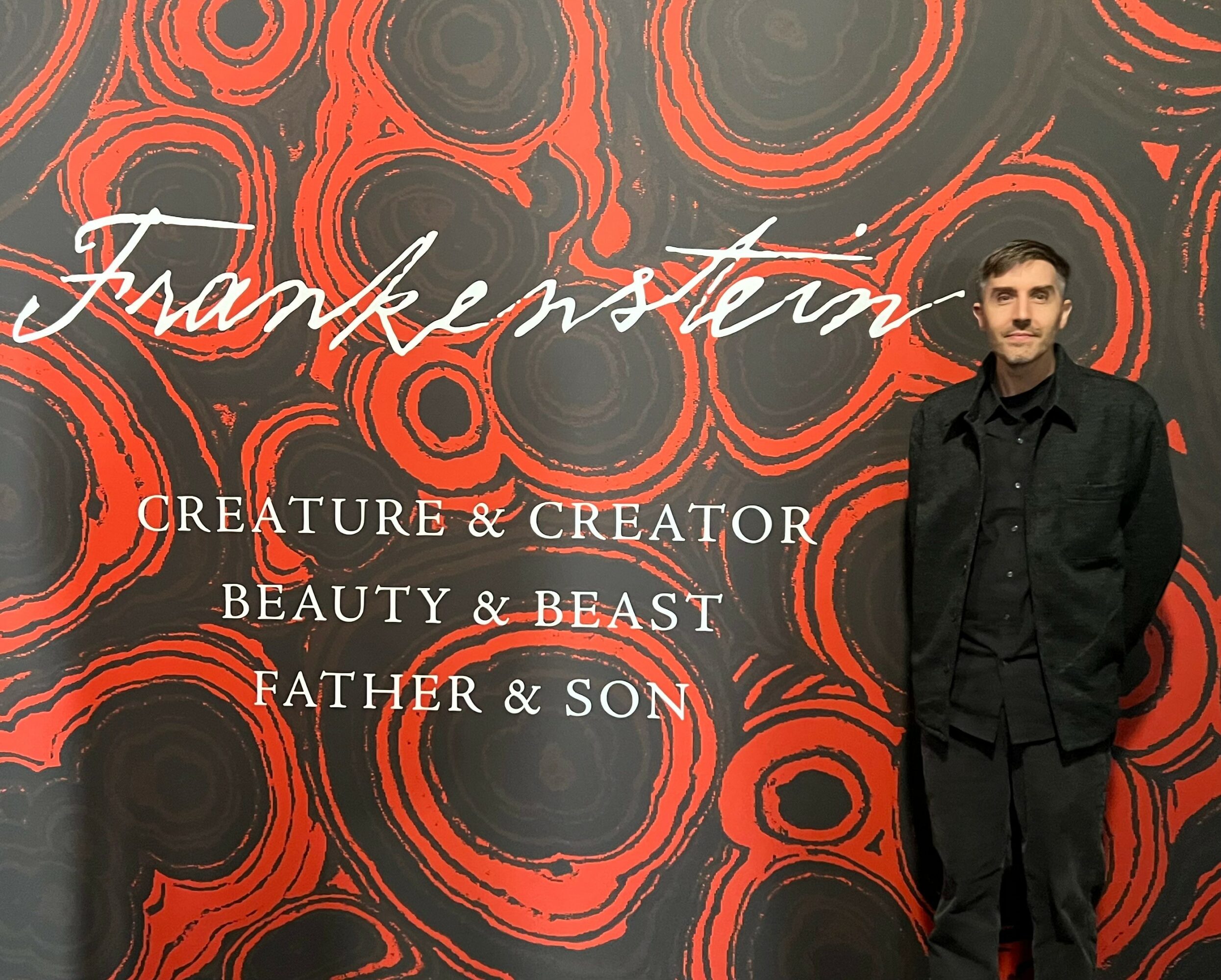 Man in black coat standing in front of a red and black abstract background with text.