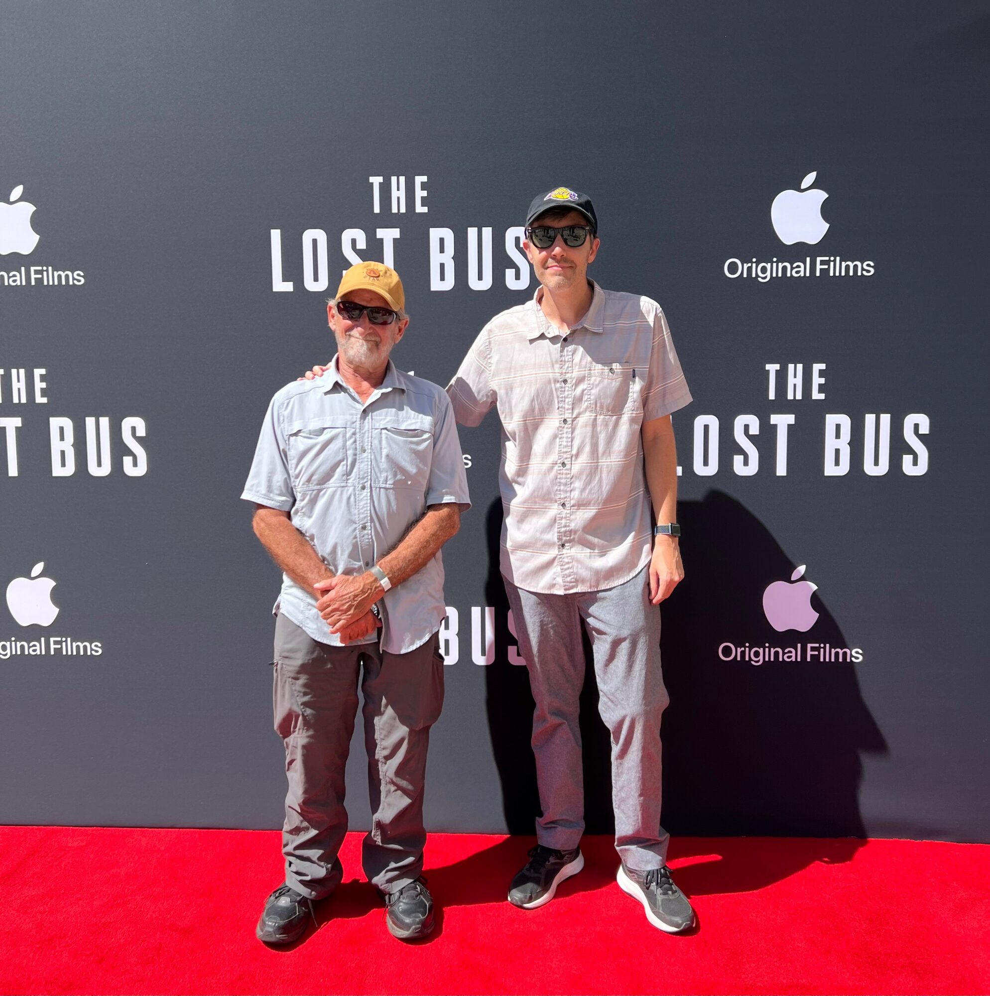 Two men standing on a red carpet in front of a black backdrop with logos and text, smiling at the camera.