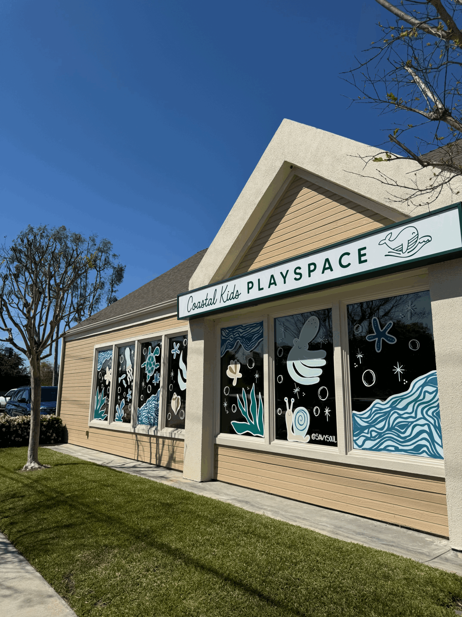 Building with large windows displaying colorful artwork, sign reads 'PLAYSPACE,' clear blue sky, trees, and grass in foreground.
