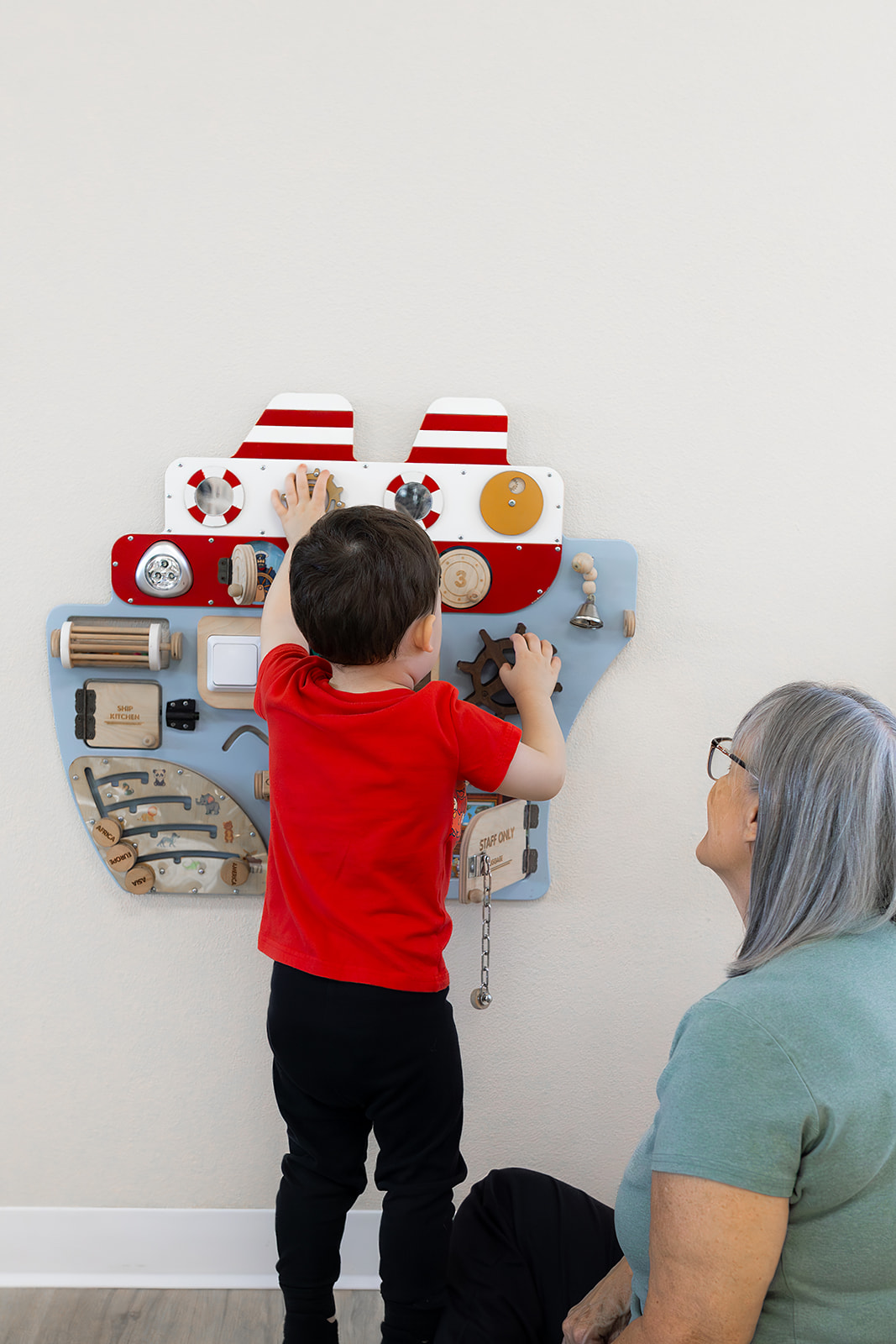 Child in red shirt playing with a wall-mounted musical toy, woman watching nearby, plain wall background.