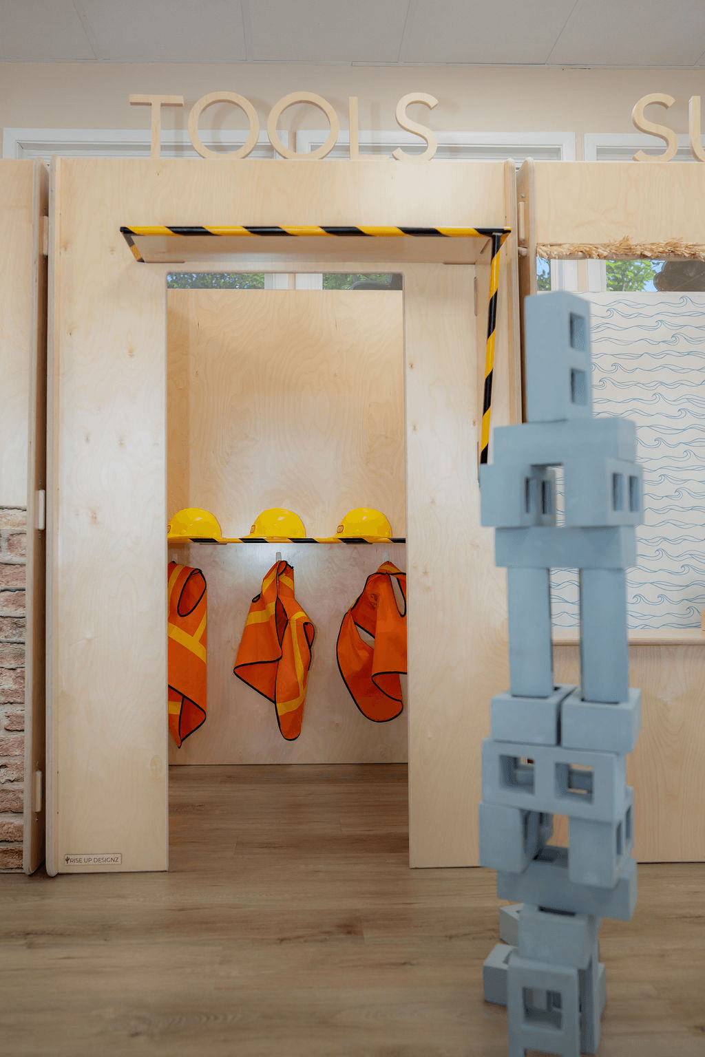 Play area with construction tools theme, yellow helmets, orange vests, and blue foam blocks, with a doorway and partial wall.