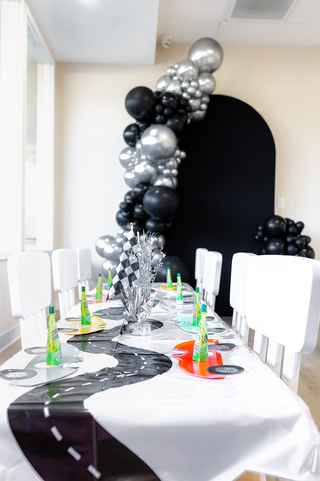 Party table with balloons, colorful party hats, and a black backdrop, decorated for celebration, with white chairs around the table.