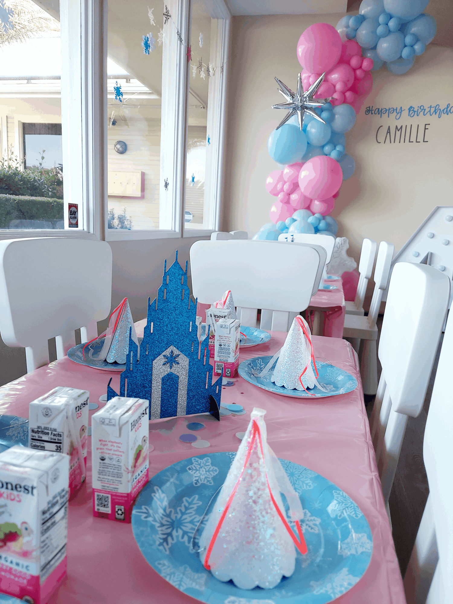 Party table with blue and pink decorations, a castle centerpiece, and balloons in a room with large windows.