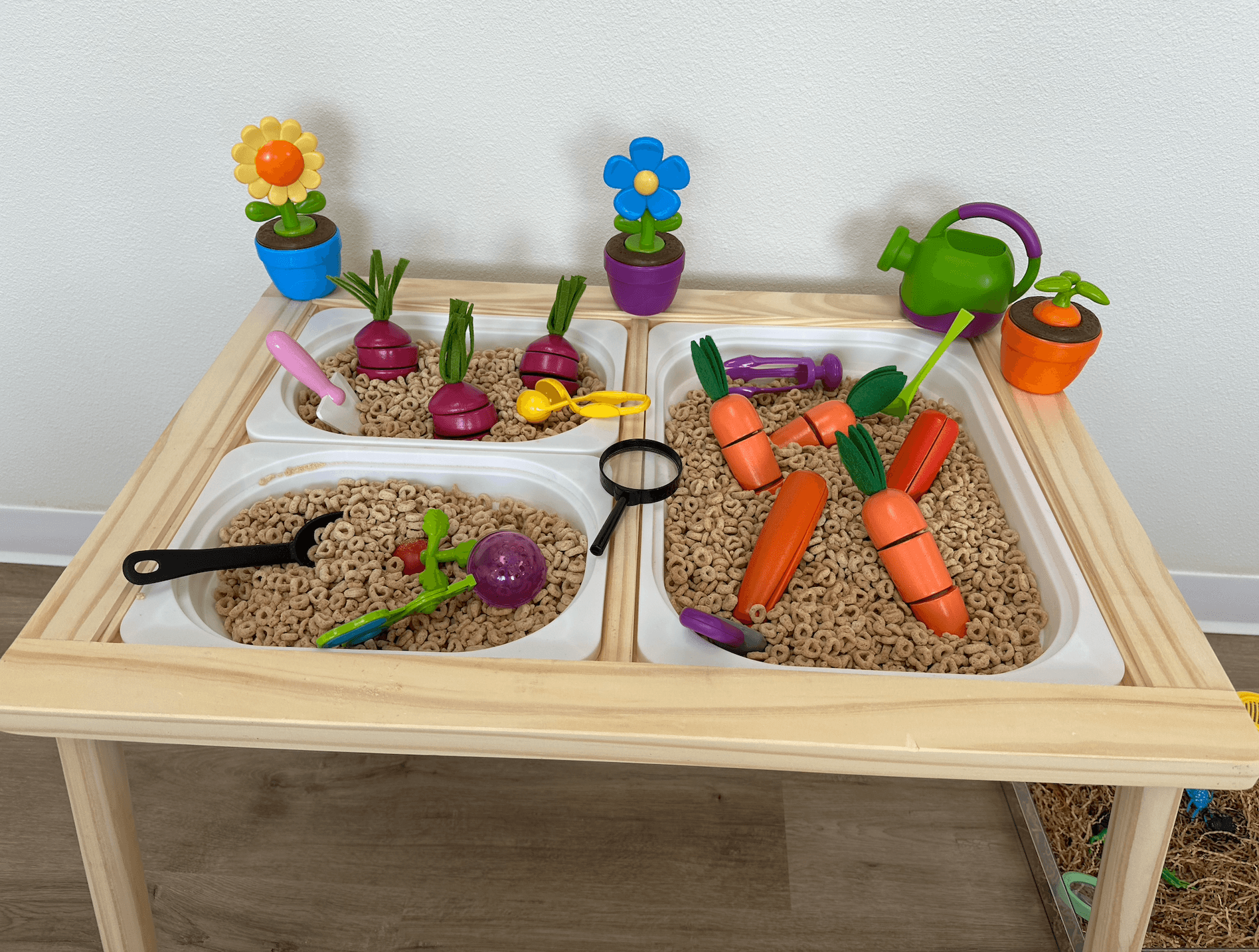 Children's play table with toy vegetables, watering cans, and flower pots on a wooden surface against a white wall.