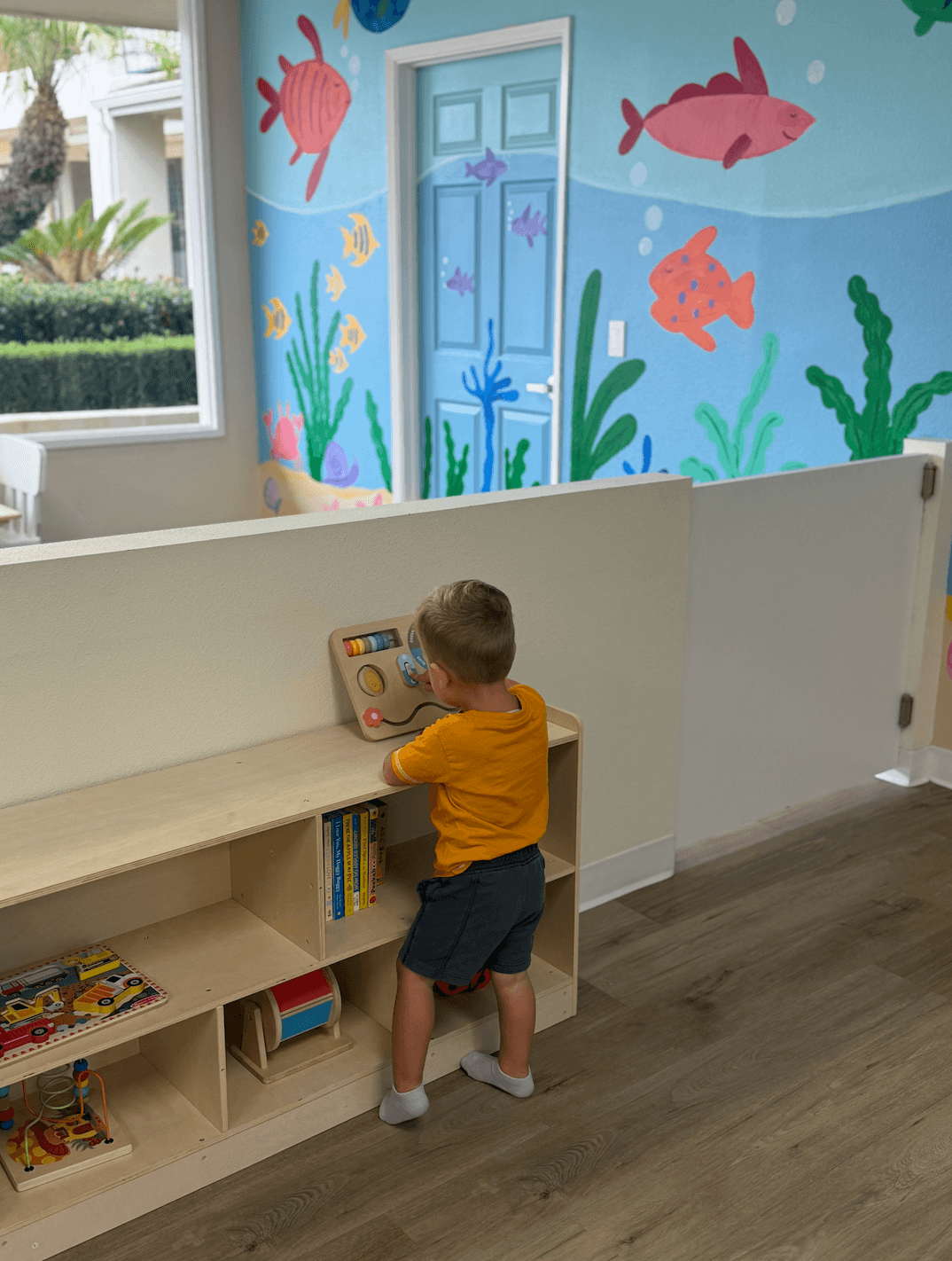Child in yellow shirt and shorts playing with a toy on a wooden shelf in a colorful room with ocean-themed wall art.