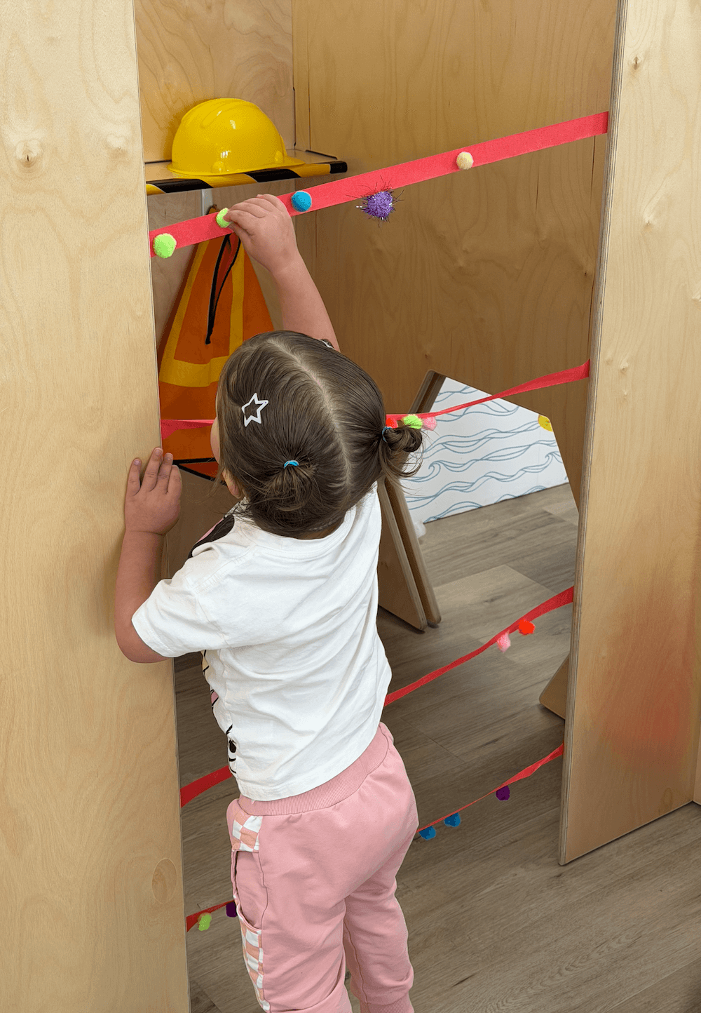Child with a ponytail and hair clips reaching for a yellow helmet on a wooden structure with colorful balls and ribbons, indoors.