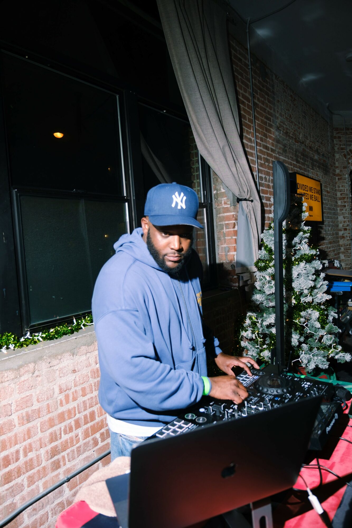 Person wearing a blue hoodie and a blue New York Yankees cap operating DJ equipment indoors, brick wall background, flowers nearby.