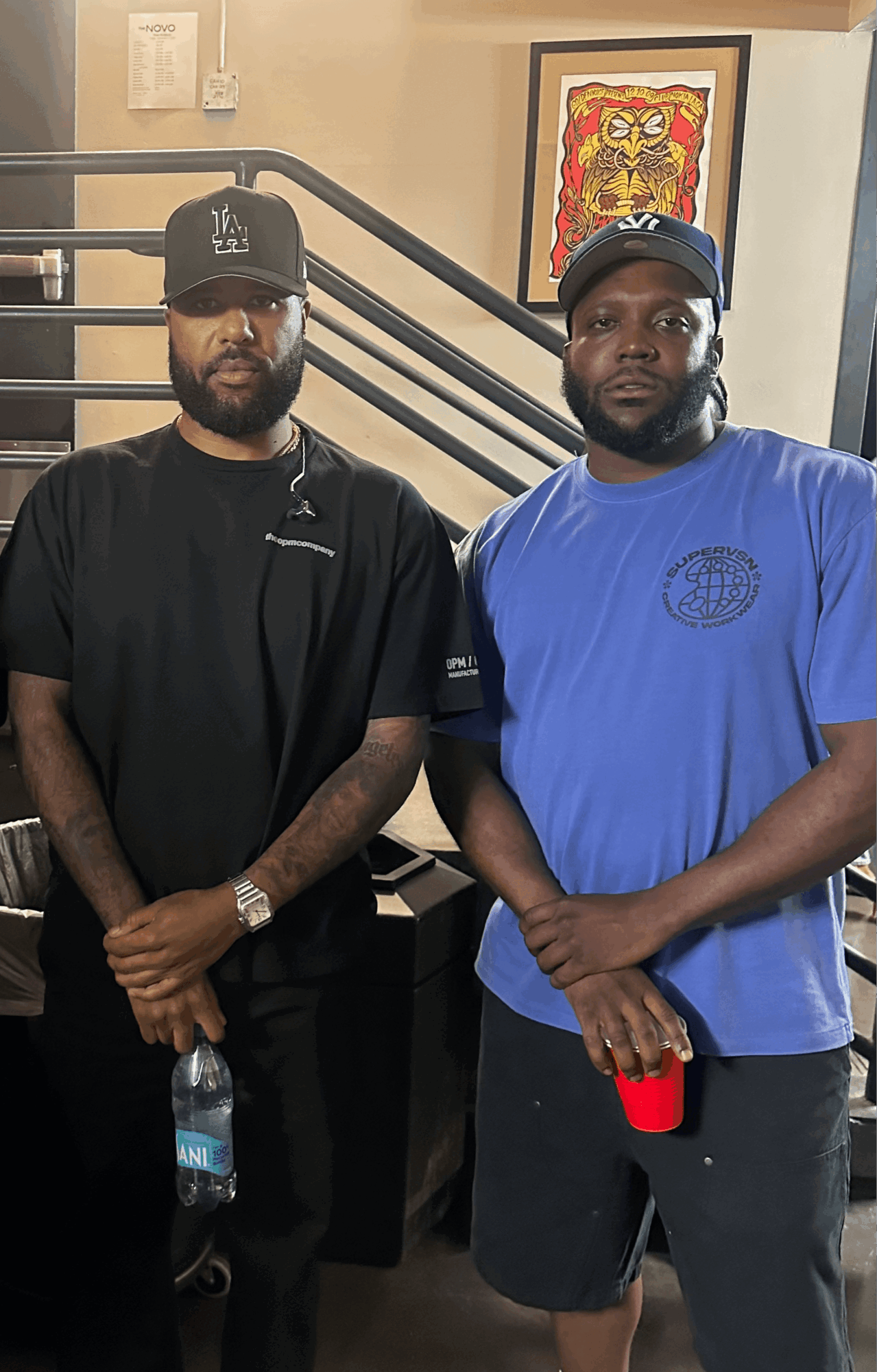 Two men stand indoors, one in black clothing and cap, the other in blue T-shirt and cap, with artwork behind them.
