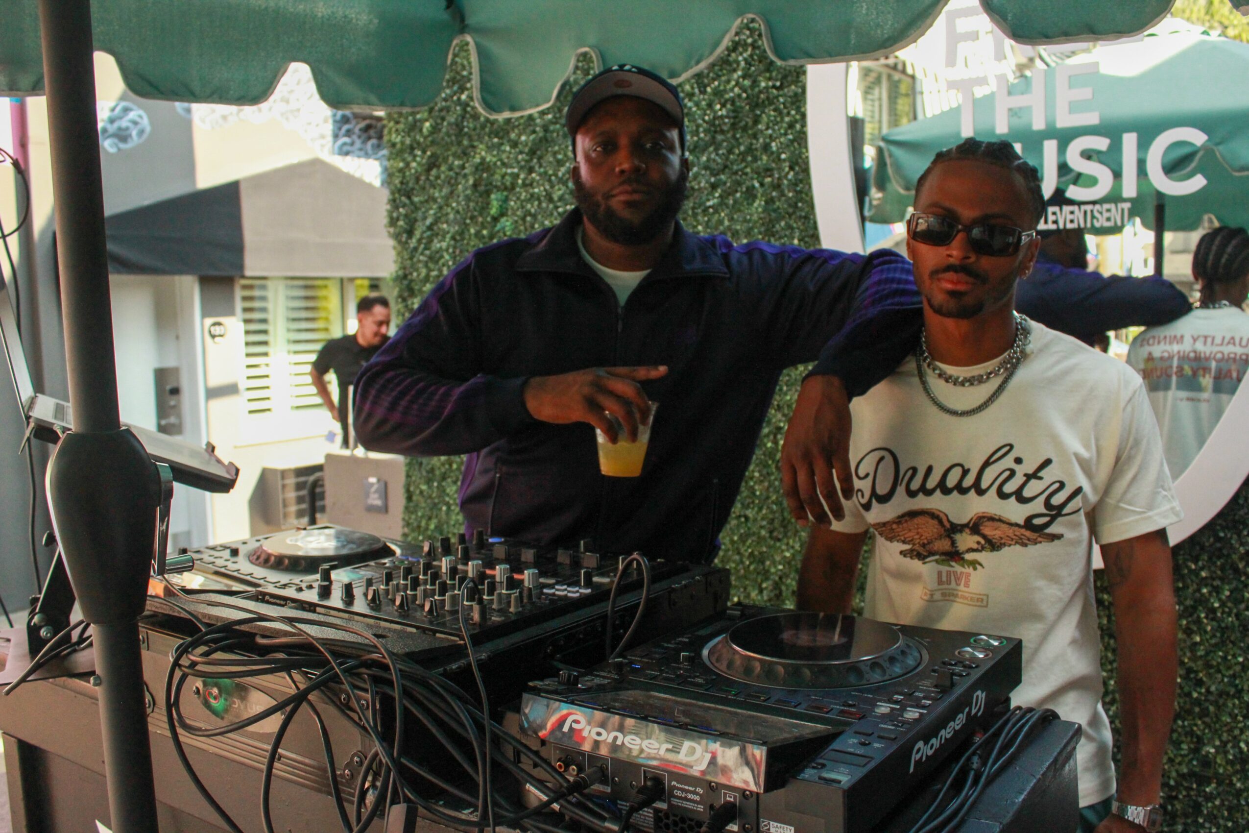 Two men standing behind DJ equipment, one holding a drink, outdoors with a green canopy and people in background.