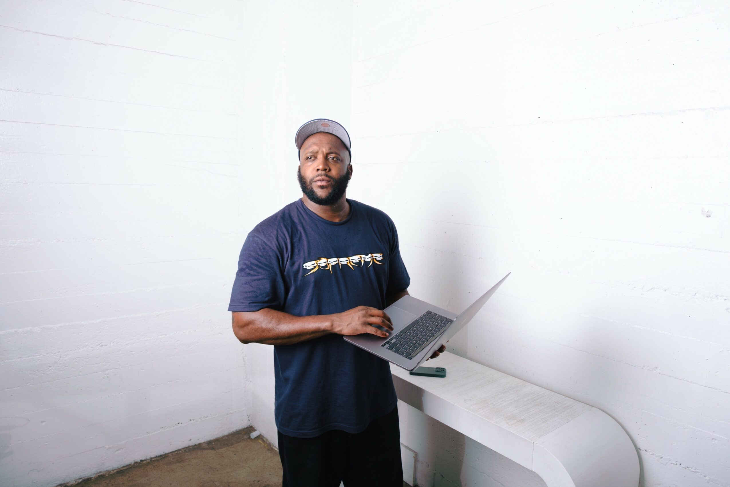 Man with a beard wearing a cap and navy T-shirt holding a laptop, standing against a white wall.