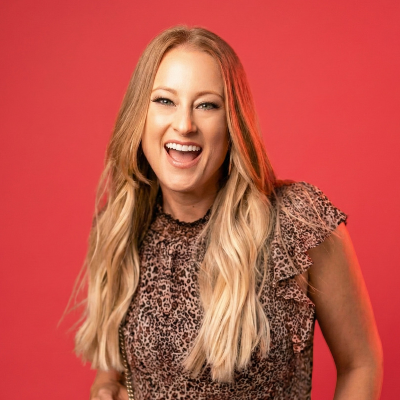 Smiling woman with long blonde hair wearing a leopard print top against a pink background.