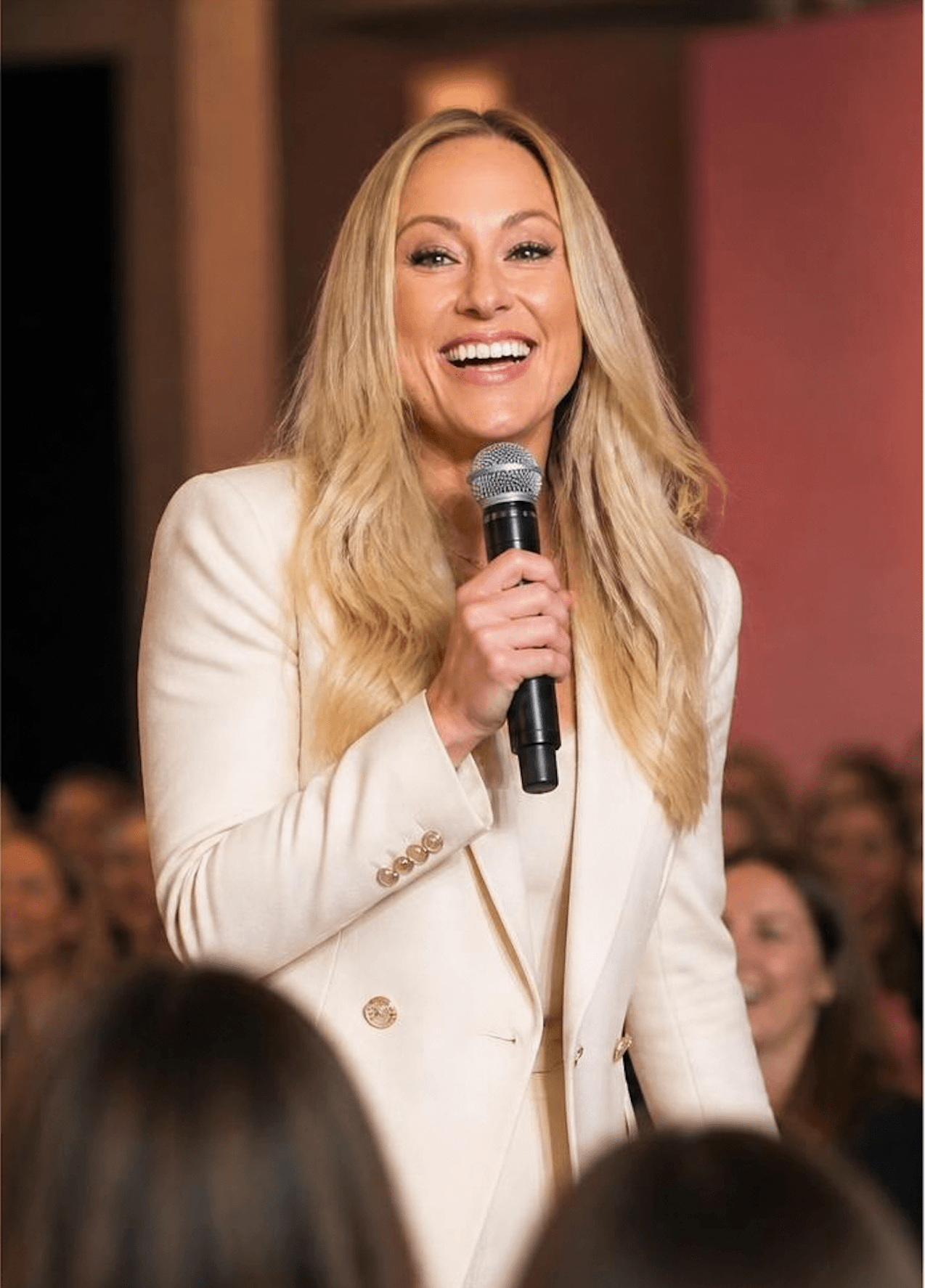 Woman with long blonde hair smiling and holding a microphone, wearing a white blazer, in front of an audience.