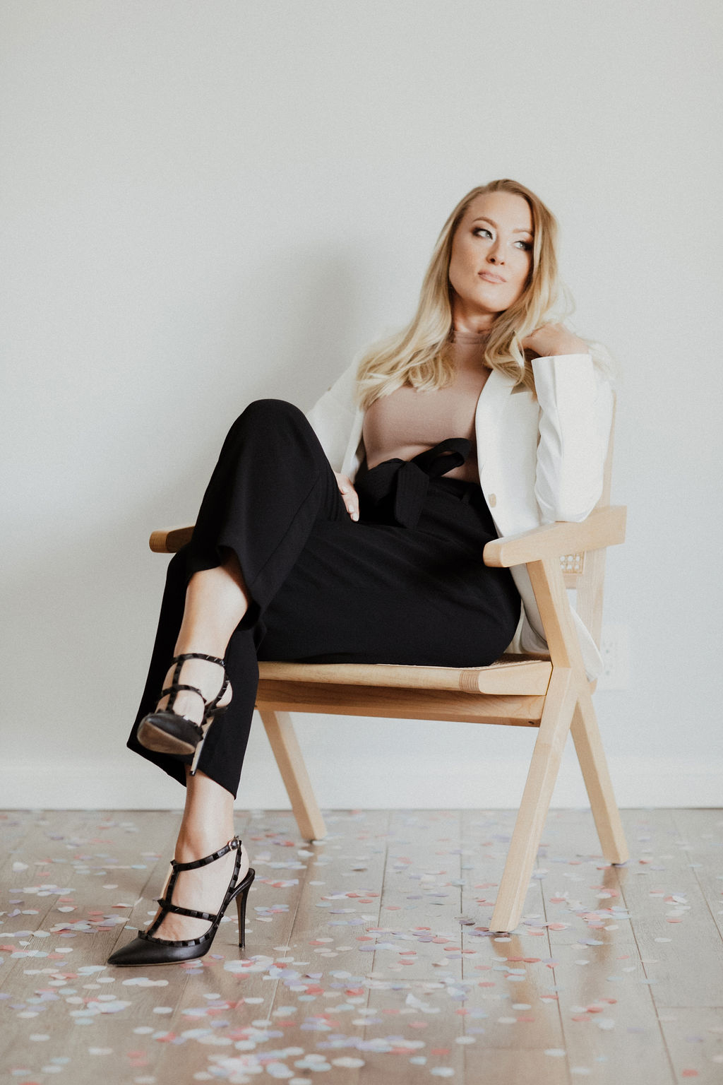 Woman sitting on a wooden chair with crossed legs, wearing a white blazer, black pants, and high heels, against a plain wall.