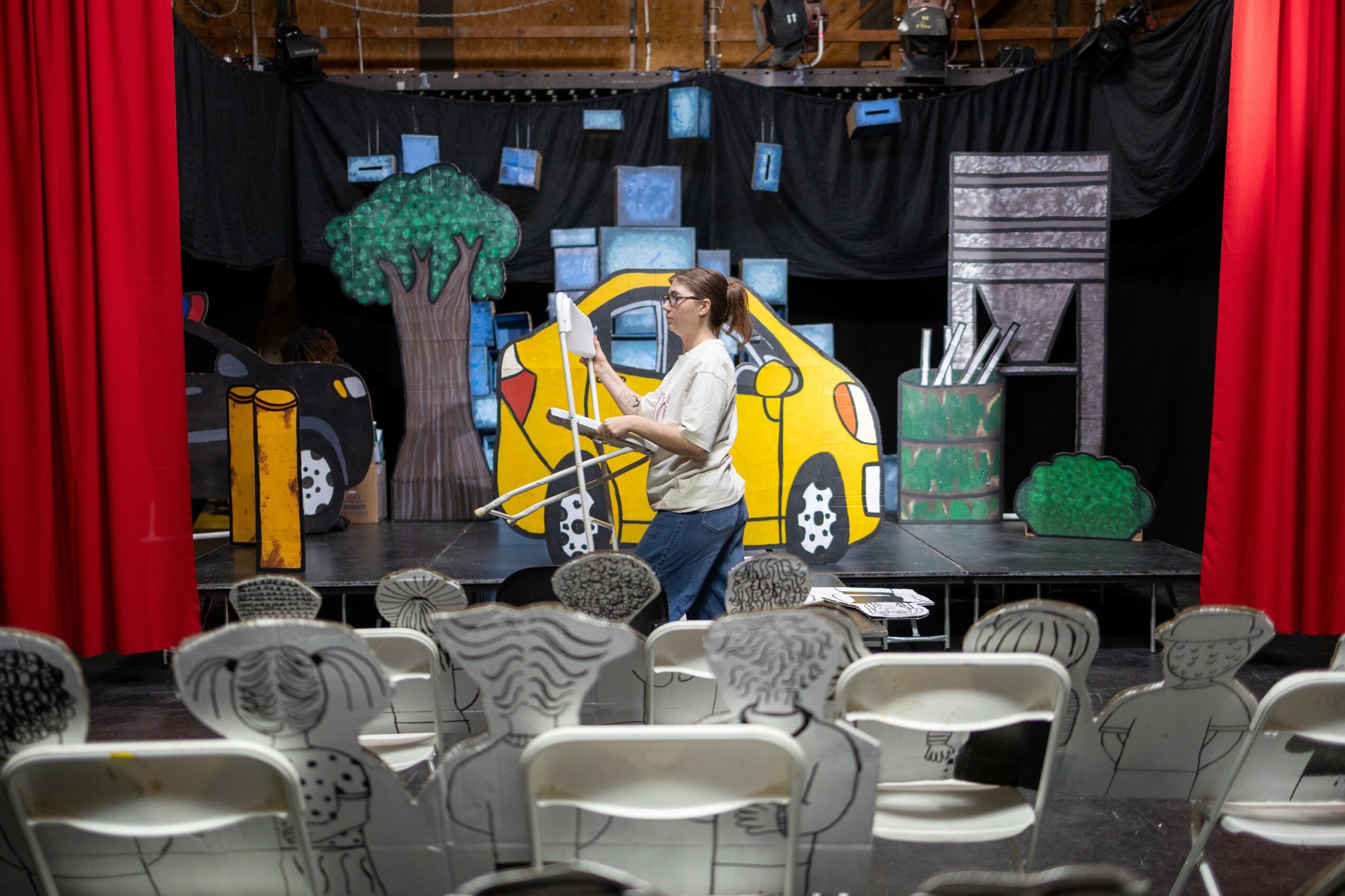Person standing on stage with colorful set pieces, chairs in foreground, red curtains on sides, black backdrop, and stage props.