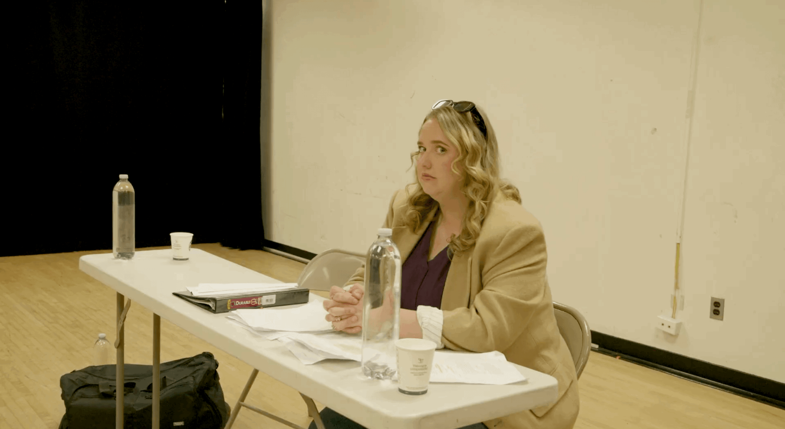 Woman sitting at a table with water bottle, papers, and a cup, in a room with a whiteboard.