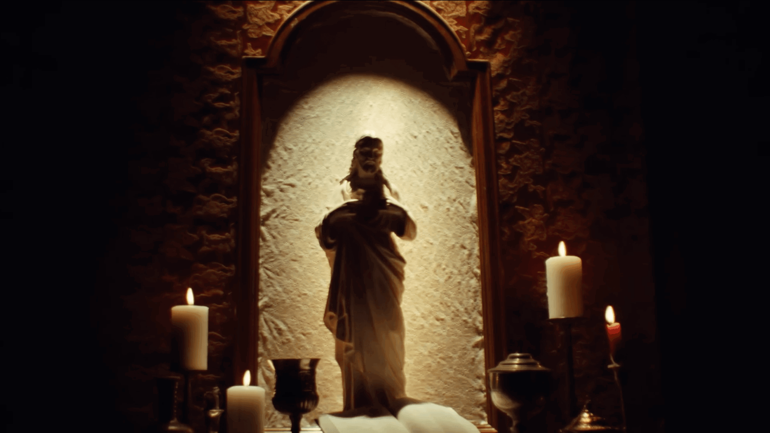 Statue of a person holding an object, illuminated by a spotlight, surrounded by candles on a dark background.