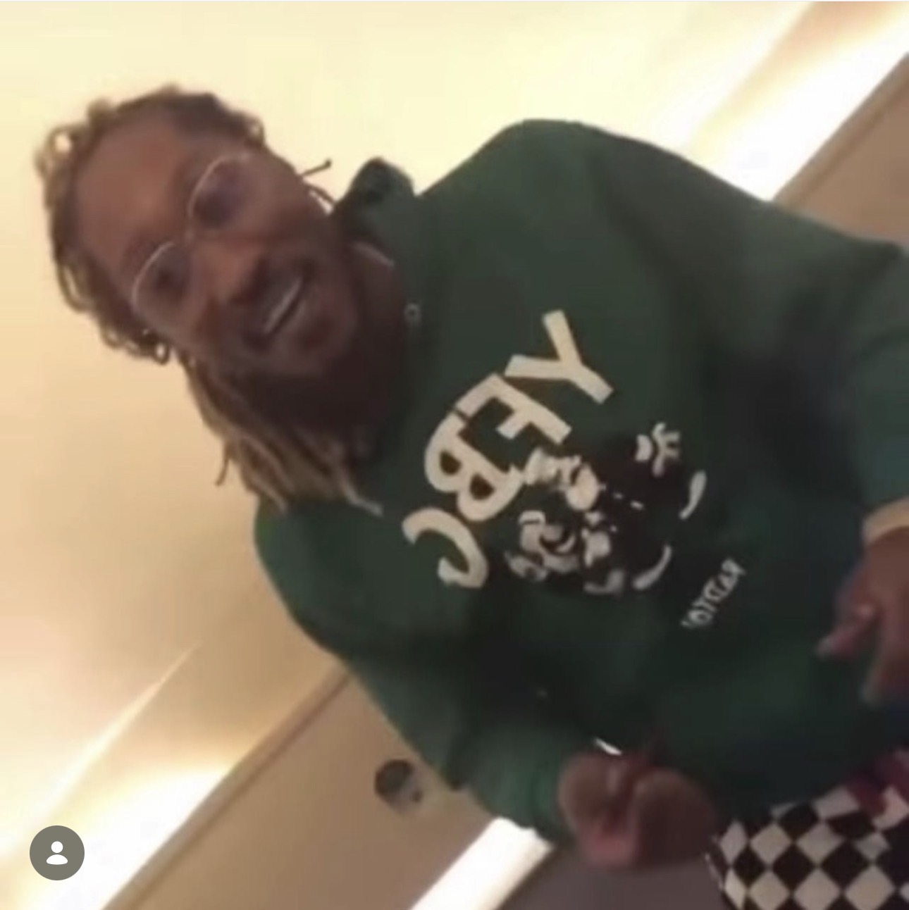 Person with glasses and dreadlocks smiling, wearing a green hoodie with white text, indoors.