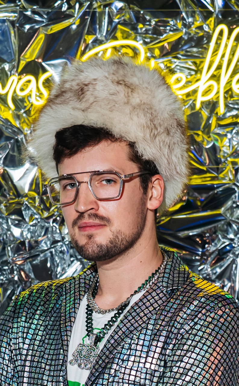 Young man with glasses wearing a furry hat and a shiny, patterned jacket, standing in front of a metallic background.